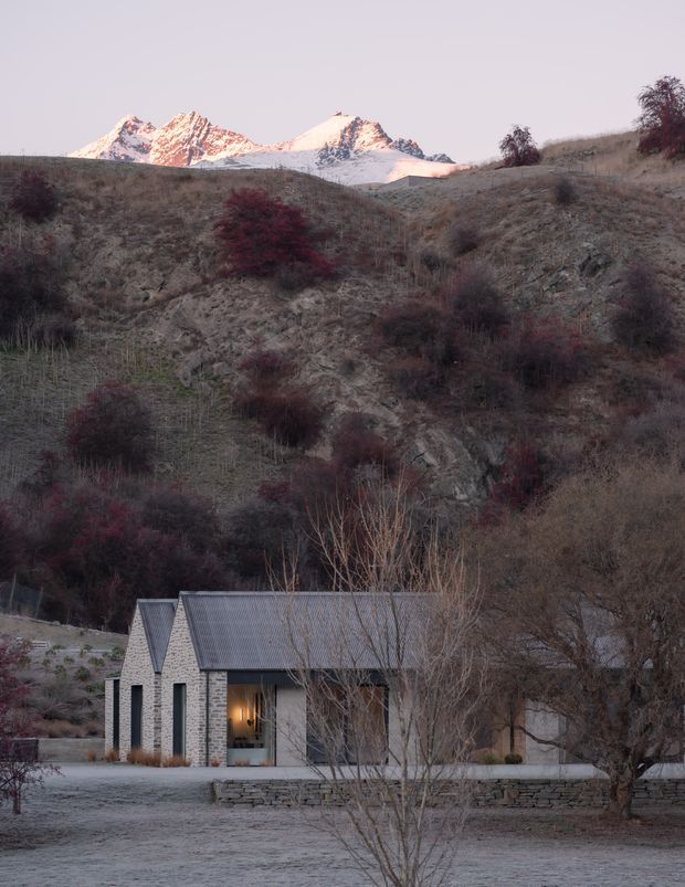 A small single-storey stone-clad dwelling sits in a rugged alpine landscape beneath snow-capped mountains. The building has two steeply pitched gable volumes with dark corrugated metal roofing, narrow dark-framed openings, and a sheltered glazed entrance. Dry grasses, scrub, stone retaining walls, and leafless trees surround the house, creating a quiet, remote mountain setting at dusk.