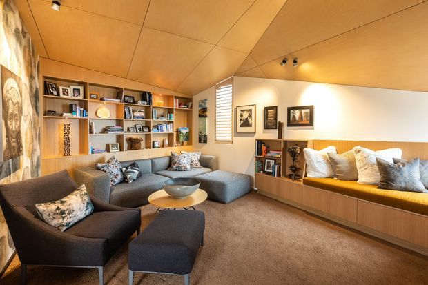 Warm, inviting mid-century-inspired living room with extensive built-in timber shelving, a low timber-paneled ceiling, integrated window seating, and a large grey sectional sofa. The room includes framed artwork, books, decorative objects, and a dark lounge chair and ottoman arranged around a small round coffee table. A textured stone or exposed masonry feature wall is visible at left.