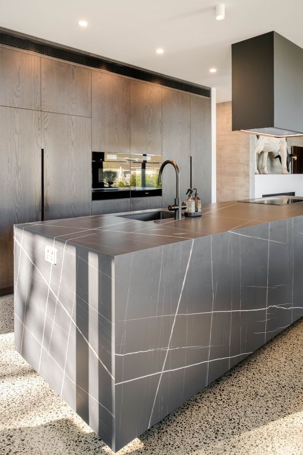 A contemporary kitchen dominated by a large charcoal-grey stone-clad island with prominent white veining, integrated sink and curved black tap. Full-height dark timber cabinetry lines the rear wall, incorporating black appliances and a concealed linear air vent. A large dark rangehood hangs above the cooking area, while terrazzo flooring, recessed ceiling lights, and warm timber surfaces create a refined, high-contrast fit-out.