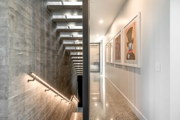 A contemporary interior hallway beside a dramatic concrete stairwell. The stair descends along a textured raw concrete wall with a slim illuminated handrail and exposed timber-and-metal treads beneath a linear skylight or clerestory. The adjacent corridor has white walls, recessed ceiling lights, polished aggregate or terrazzo flooring, and a series of framed decorative panels.