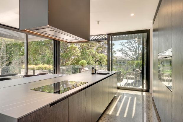 Contemporary kitchen with a large island, integrated cooktop, undermount sink, dark linear-grain cabinetry, and pale stone benchtops. Floor-to-ceiling glazing and sliding doors open to a bright outdoor setting, while a sculptural rangehood forms a strong ceiling feature. The room has a refined, airy and highly integrated feel.
