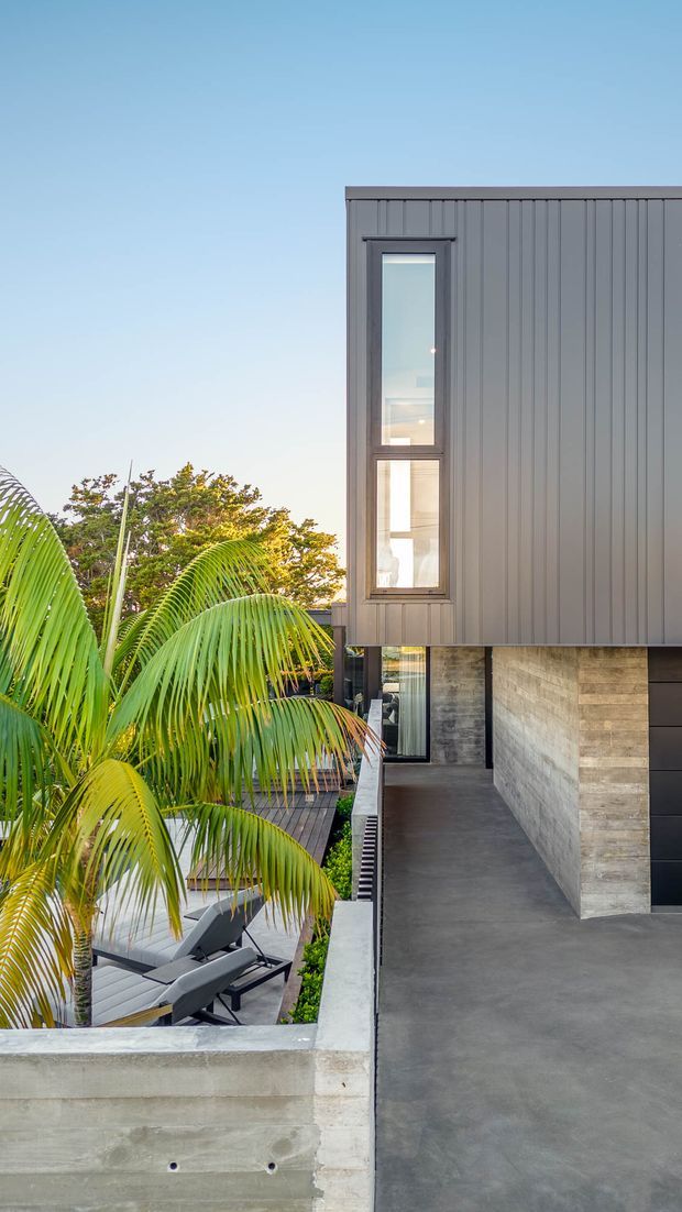 Exterior view of a contemporary multi-level residence with dark vertically ribbed metal cladding, narrow vertical windows, exposed concrete outdoor surfaces, and a stone-clad wall. A tropical palm-filled terrace with sun loungers is visible beside the building.