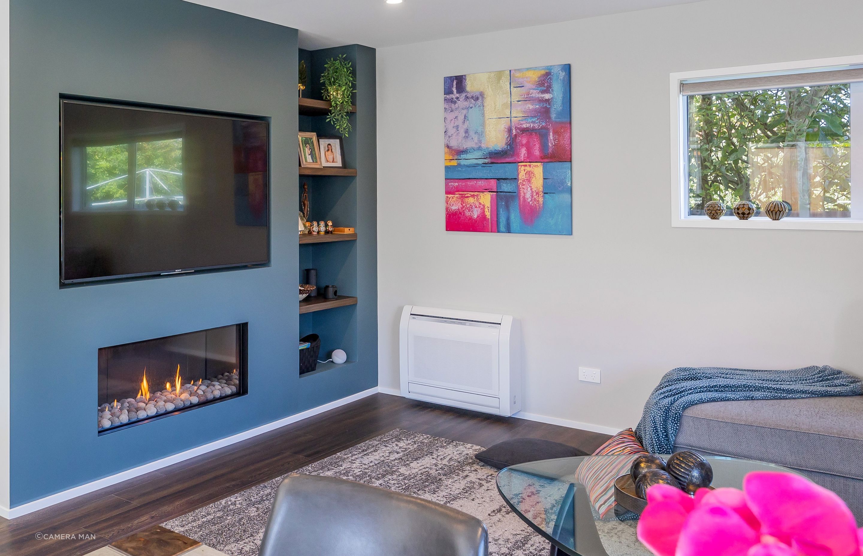 The Feature wall separating the staircase from the Living Room is a wall that was specifically built to house the TV and the Fireplace, as well as provide a recessed shelf where the client's can display photographs and personal belongings. This Living Room being part of the open plan kitchen / dining / living with access to the deck and outdoor dining, serves as a central gathering space for family members and guests to socialize, relax, and spend quality time together. It is the space where they can engage in conversations, watch movies or TV shows, play games, and enjoy various forms of entertainment. This living room acts as the focal point for hosting guests and creating a welcoming atmosphere.