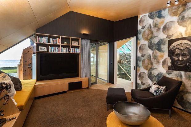 A warm, compact contemporary living room with a built-in timber media wall and open shelving, dark charcoal feature walls, timber ceiling, and brown carpet. A mustard built-in bench sits beside the television, while two dark lounge chairs and a round timber coffee table face the media unit. Floor-to-ceiling glazing and a glazed door connect the room to an exterior stair and coastal outlook. One wall features a bold decorative mural with a sculptural face relief.