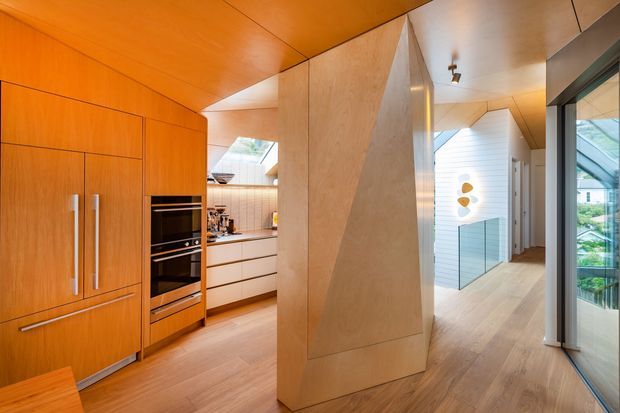 Warm contemporary open-plan interior showing a compact kitchen and adjoining hallway. The space features extensive honey-toned timber cabinetry and ceiling panels, integrated stainless-steel appliances, pale timber flooring, sculptural angled timber wall panels, and glazing opening toward an exterior garden or courtyard.