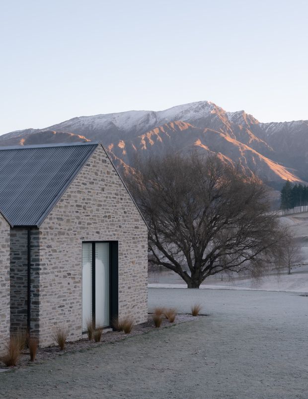 Exterior view of a compact stone-clad gable-roof building in a frosty alpine landscape. The building has pale masonry walls, a dark corrugated metal roof, and a tall narrow glazed door or window with dark framing. A mature leafless tree and rugged snow-dusted mountains form the background.