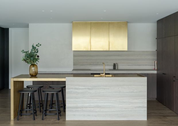 Contemporary minimalist kitchen with a large stone-clad island, integrated timber breakfast bar and black stools. The room features dark timber floor-to-ceiling cabinetry, pale horizontally grained splashback panels, brass-toned upper cabinetry, recessed ceiling lights, and a muted neutral palette.