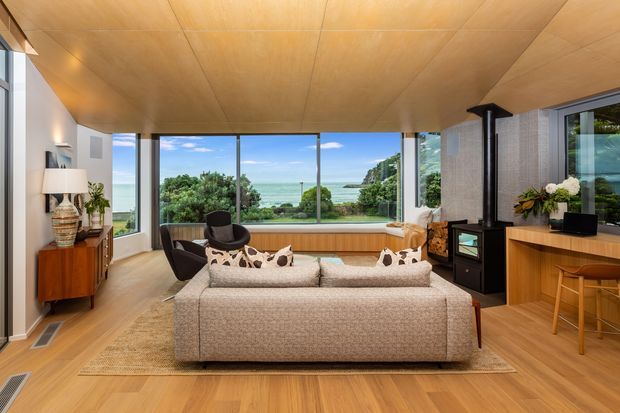 A warm, timber-lined living room with expansive floor-to-ceiling glazing overlooking a lush coastal landscape and water. The room features a large neutral sectional sofa, mid-century-inspired lounge chair, timber flooring and cabinetry, a black freestanding wood-burning stove, and a compact timber bar or kitchenette area.