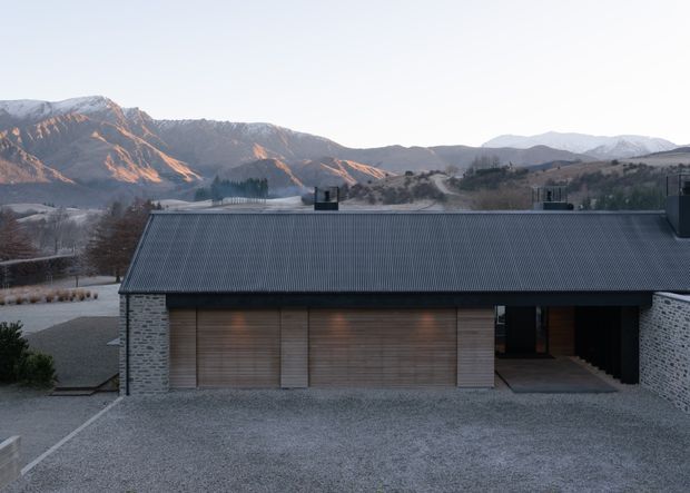 A low, modern building viewed from outside, set against a dramatic mountain landscape. The facade combines pale stacked-stone walls with warm timber garage doors and a dark corrugated pitched roof. A broad gravel forecourt and recessed entry create a restrained, contemporary composition.