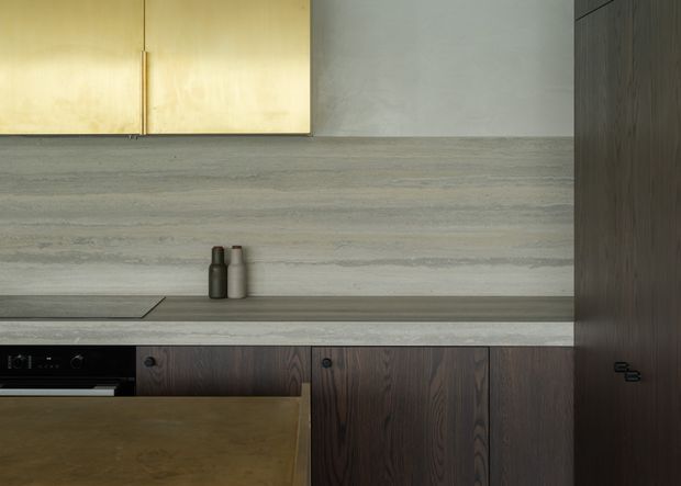 Close-up of a refined contemporary kitchen with dark timber cabinetry, a long grey stone benchtop and matching full-height stone splashback. Brass-toned upper cabinet fronts provide a warm metallic accent, while integrated appliances and minimal hardware create a clean, understated composition.
