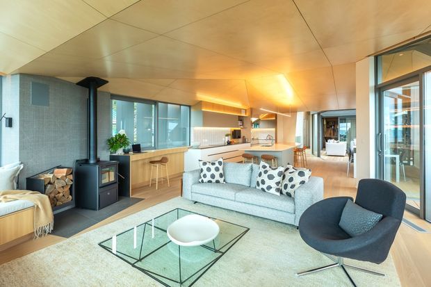Open-plan contemporary living space with a lounge, kitchen and dining area. The room features extensive glazing, warm timber ceiling and flooring, a pale upholstered sofa, sculptural dark lounge chair, glass coffee table, and a black freestanding wood-burning stove beside a tiled feature wall and firewood storage.