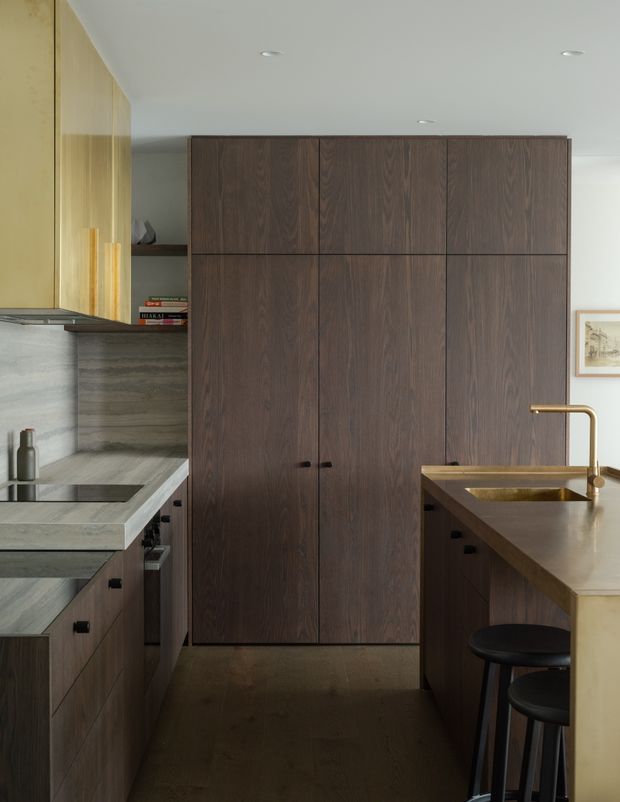 A refined compact kitchen with dark timber cabinetry, tall integrated storage, grey stone benchtops and splashback, and a warm metallic tap over a small undermount sink. A peninsula with bar stools occupies the right side, while pale timber wall panelling and recessed ceiling lights add warmth. The space has a restrained, high-end contemporary appearance with a narrow galley-like layout.