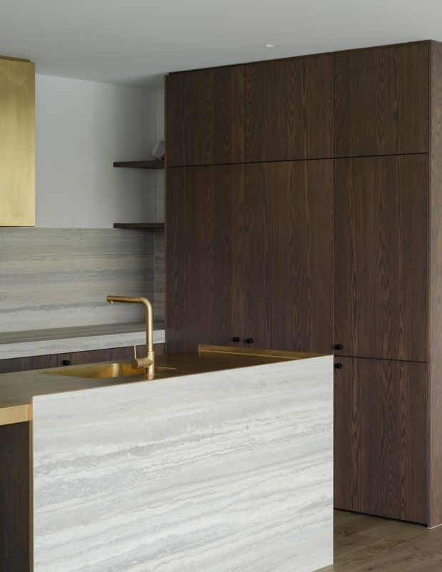 A refined contemporary kitchen featuring a substantial pale veined stone island and matching rear benchtop, dark walnut-toned full-height cabinetry, and a brushed brass gooseneck tap with integrated sink. Concrete-look or stone wall and flooring surfaces create a restrained, luxurious material palette. A stair or stepped architectural element is visible in the background.