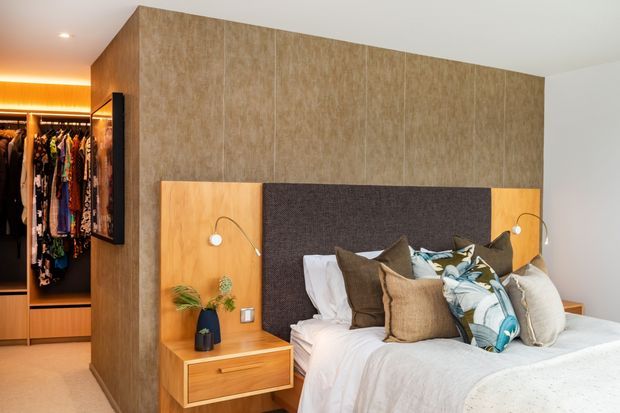A contemporary bedroom with a large upholstered headboard, integrated timber bedside niches and drawers, wall-mounted reading lights, layered neutral bedding, and a walk-in wardrobe visible through the opening at left. The room features warm timber-look wall panelling and softly concealed lighting.