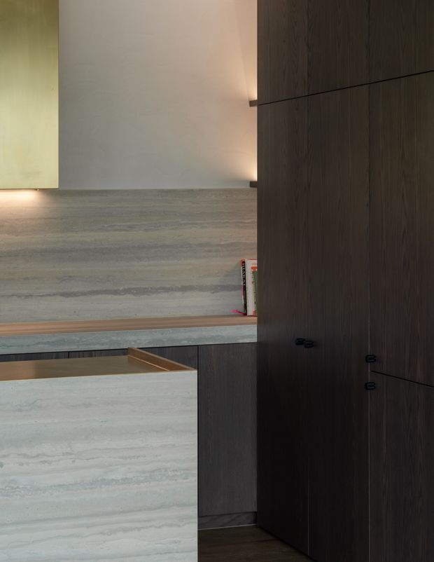 A close-up architectural interior detail showing dark timber built-in cabinetry beside a pale, horizontally veined stone-clad wall and island or counter. Recessed warm lighting creates a refined, minimalist atmosphere, with a small open shelf containing books and a glimpse of a timber-lined lower surface.