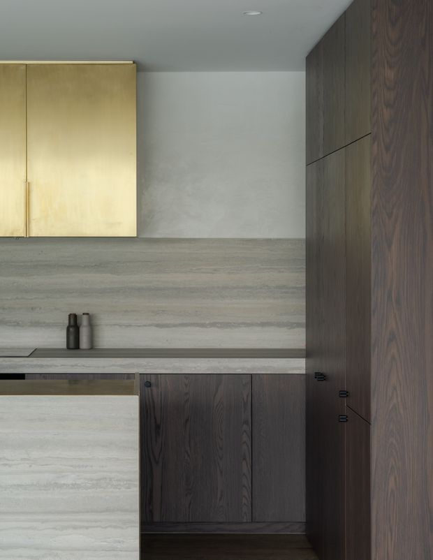 A close-up view of a minimalist kitchen featuring dark timber cabinetry, pale brushed or honed stone surfaces, a matching stone backsplash and benchtop, and a small integrated cooktop area. Brass-toned upper cabinet fronts contrast with the dark wood joinery. The restrained palette and concealed hardware create a refined, contemporary interior.