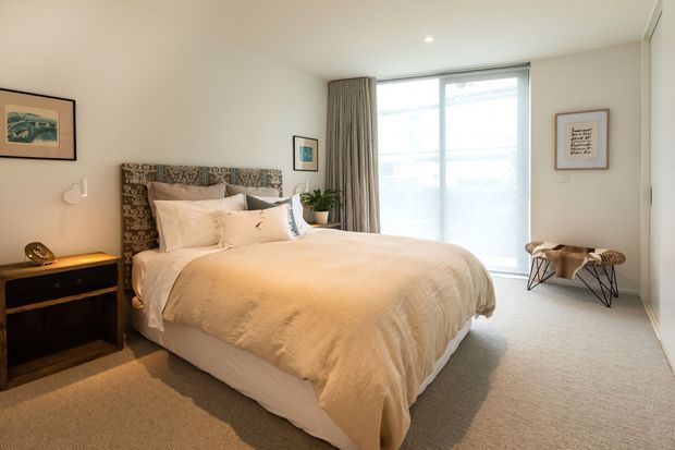 Bright, softly styled bedroom with a large upholstered bed, patterned headboard, cream bedding, beige carpet, dark bedside furniture, framed artwork, a plant, and full-height window glazing with curtains.