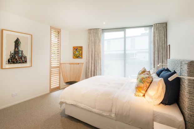 Bright, softly styled bedroom with a large upholstered bed, white bedding, colourful accent cushions, woven bedside furniture, full-height curtained glazing, timber shutters, pale carpet, and neutral walls. Artwork and a sculptural timber console add restrained decorative detail.