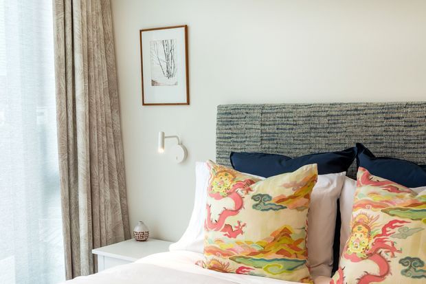 A softly lit bedroom featuring a large upholstered textured headboard, layered white bedding, dark navy pillows, and colourful patterned accent cushions. Floor-to-ceiling textured curtains frame a bright window, while a minimal wall sconce and small framed artwork add understated detail.