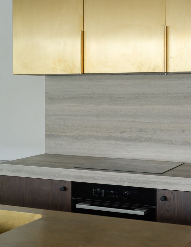 Close-up view of a contemporary kitchen cooking zone, featuring pale timber overhead cabinetry, a horizontally veined grey stone backsplash, integrated dark cabinetry, a black induction cooktop, and a built-in black oven. A muted stone or concrete-look benchtop is visible in the foreground.