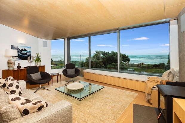 A bright, refined living room with expansive floor-to-ceiling glazing overlooking the ocean. The room features a warm timber ceiling, white walls, a built-in timber window seat or low cabinet, neutral carpet, a glass coffee table, sculptural lounge chairs, a pale sectional sofa, and a compact desk area. The coastal outlook and generous glazing create an airy, relaxed atmosphere.