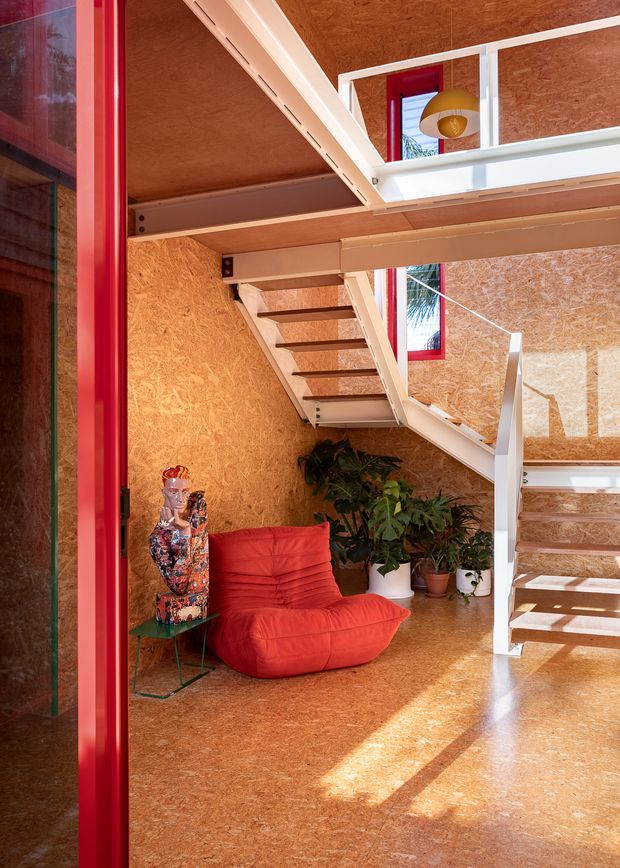 A striking multi-level interior with exposed OSB-style timber wall, floor and ceiling surfaces, white-painted steel or timber stair structures, and vivid red window and door frames. A sculptural red lounge chair sits beside potted plants and a decorative mannequin, while an upper-level landing with white balustrading and a pendant light overlooks the space. Strong sunlight creates bold shadows across the floor.