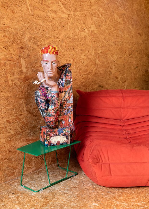 A vivid, eclectic interior vignette featuring a red upholstered lounge chair or compact sofa, a colourful mannequin sculpture, and a small green metal side table. The walls and floor are clad in exposed oriented strand board (OSB), creating a strongly textured, warm-toned backdrop. The composition has a playful, bold and retro-pop character.
