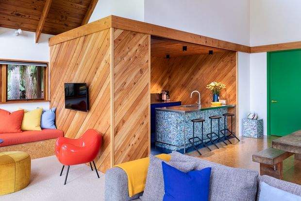 Vibrant open-plan living and entertaining space with a built-in timber-lined partition and compact kitchen/bar. The room features a warm exposed timber ceiling, diagonal timber wall panelling, colourful upholstered seating, a patterned blue mosaic island, bar stools, and a bright green internal door.