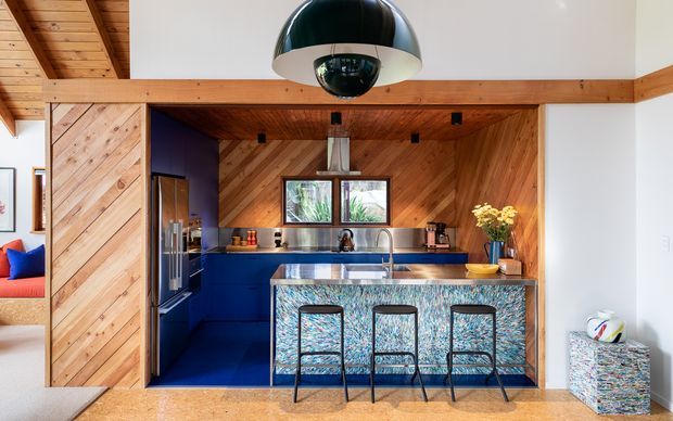 A vibrant open-plan kitchen with a deep blue island and cabinetry, three bar stools, and a highly patterned stone or terrazzo island face. Warm diagonal timber panelling covers the walls and ceiling, contrasted with white walls, stainless-steel appliances, and a large black-and-chrome dome pendant light. A living area is partially visible at left, while a small colourful upholstered stool sits at right.
