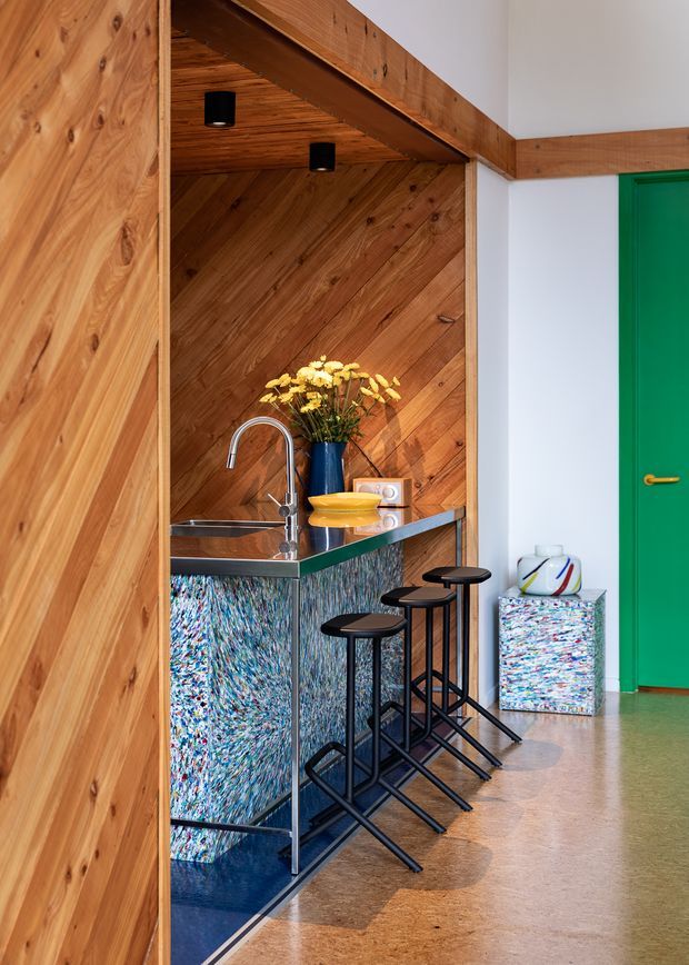 A compact, colourful home bar or kitchenette set into a timber-lined alcove. The space features a stainless-steel sink and tap, glossy blue reflective cabinetry with multicoloured textured fronts, a stone or composite benchtop, three black bar stools, and a bright green door. Warm timber panelling continues across the walls and angled ceiling, creating a bold contemporary interior.