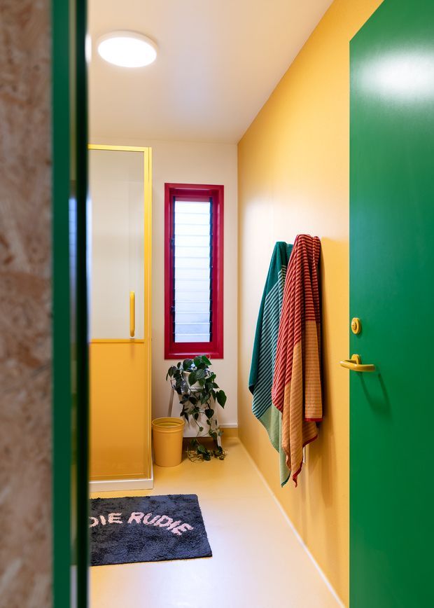 A brightly coloured compact bathroom viewed through an open green door. The room features yellow walls and floor, a red-framed louvred window, yellow shower enclosure, colourful striped towels on wall hooks, a small potted plant, and a dark bath mat. The space has a playful, bold, coordinated colour scheme with glossy painted joinery and trim.