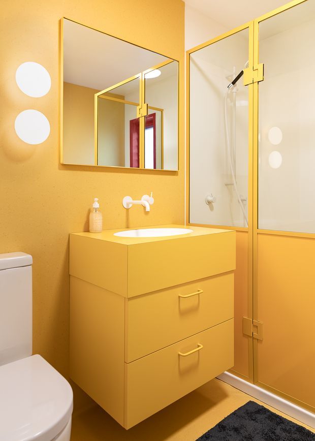 A compact, vividly monochromatic yellow bathroom with a wall-mounted two-drawer vanity, integrated white basin, wall-mounted tapware, mirrored cabinet, toilet, and framed glass shower enclosure. The shower and vanity use coordinated yellow framing and cabinetry, with white sanitaryware and a black bath mat providing contrast. Round wall lights and a reflected red-framed window add visual interest.
