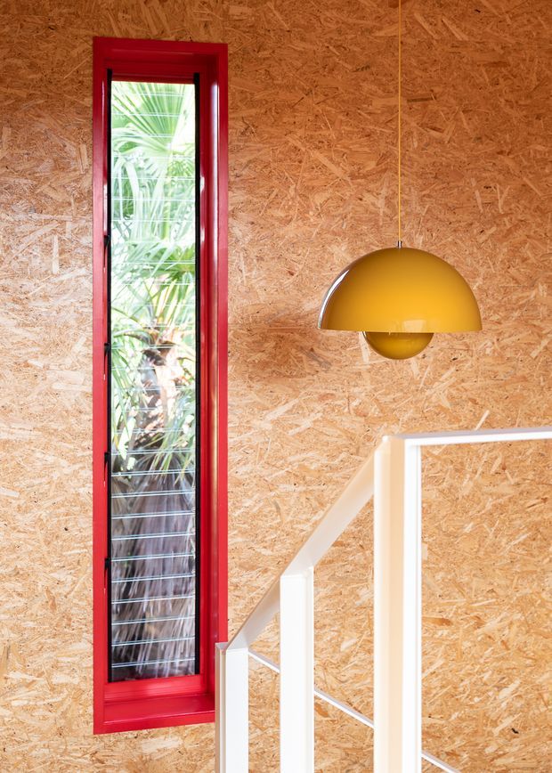 Interior architectural detail featuring an exposed oriented strand board (OSB) wall, a tall narrow window with a vivid red-painted timber frame, white stair balustrade, and a suspended mustard-yellow dome pendant light. Tropical foliage is visible through the window.