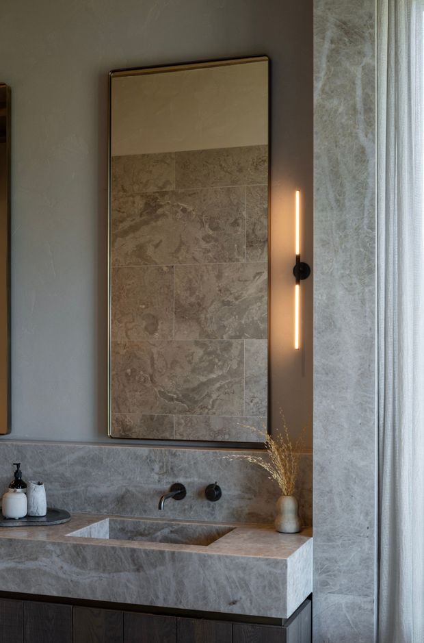 A refined contemporary bathroom vanity with a large framed mirror, grey-brown marble or stone wall and splashback surfaces, an integrated rectangular basin, dark timber cabinetry, black wall-mounted tapware, and a slim vertical wall light. A sheer curtain and stone-clad wall are visible at the right edge.