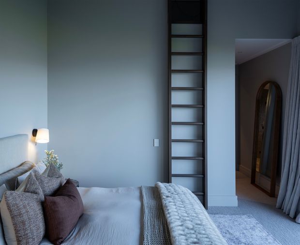 A softly lit bedroom with a pale blue-grey wall, upholstered bed, layered neutral and mauve cushions, and a chunky knitted throw. A narrow dark timber ladder-style shelving unit is built into the wall beside the bed. At right, an open doorway leads to a dim hallway or adjoining space with an arched full-length mirror, while floor-length grey curtains and a pale textured rug complete the room.