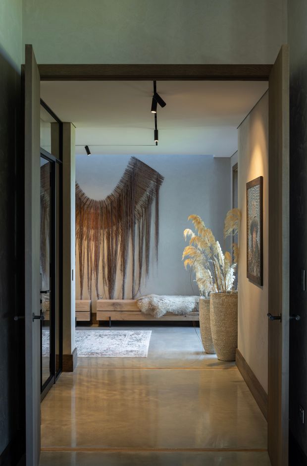 A moody, refined interior view through open double doors into a calm bedroom or sleeping nook. The space features polished concrete flooring, pale plaster walls, dark timber door framing and trim, black track lighting, a low timber platform bed or bench with textured bedding, a large organic woven wall hanging, a neutral patterned rug, framed artwork, and a tall woven planter filled with dried grasses.