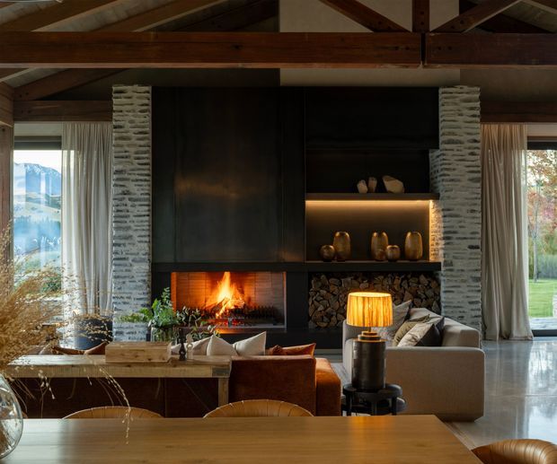 Warm, rustic-modern living room with a dramatic double-height timber-beamed ceiling and a central fireplace wall. The wall combines pale stacked-stone columns with dark built-in cabinetry, open illuminated shelving, a wood-burning fireplace, and integrated firewood storage. Large glazed openings with sheer curtains frame views of a green landscape and mountains. Furnishings include a neutral sofa, leather-toned lounge chairs, a timber coffee table, decorative objects, and a table lamp, creating a refined lodge-like atmosphere.