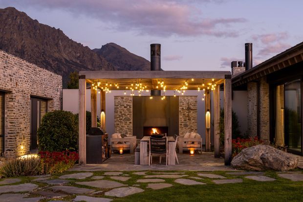 A landscaped courtyard-style outdoor lounge beside a rustic stone-clad building, set against rugged mountain terrain. The covered pergola features exposed timber posts, a flat roof, hanging string lights, a central outdoor fireplace, and lounge seating arranged around a dining table. Irregular stone paving is set into grass, with planted garden beds and warm evening lighting creating an inviting alpine retreat atmosphere.