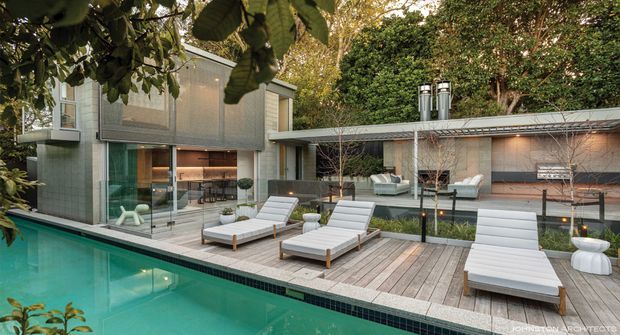 A contemporary residential pool courtyard viewed from the outdoor pool area. The scene features a turquoise swimming pool, timber decking, several white sun loungers, and a modern two-part house with extensive glazing, a flat-roofed pavilion, and landscaped screening vegetation. The architecture combines glassy upper-level volumes with a darker horizontal pavilion and outdoor entertaining area.