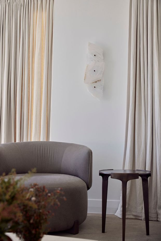 A softly lit, refined seating corner with a curved taupe upholstered armchair, a dark timber side table, full-height cream drapery, pale walls, and a small abstract wall artwork. The muted neutral palette and sculptural furniture create a calm, understated interior.