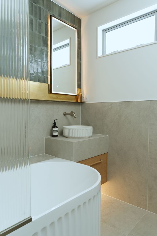 Bright contemporary bathroom featuring a freestanding white bathtub, wall-mounted brushed metal tapware, a fluted white vessel basin on a stone-look vanity, timber drawer cabinetry, large grey-beige wall and floor tiles, and a dark green tiled mirror feature wall. Ribbed translucent glazing appears at left, with a high horizontal window providing natural light.