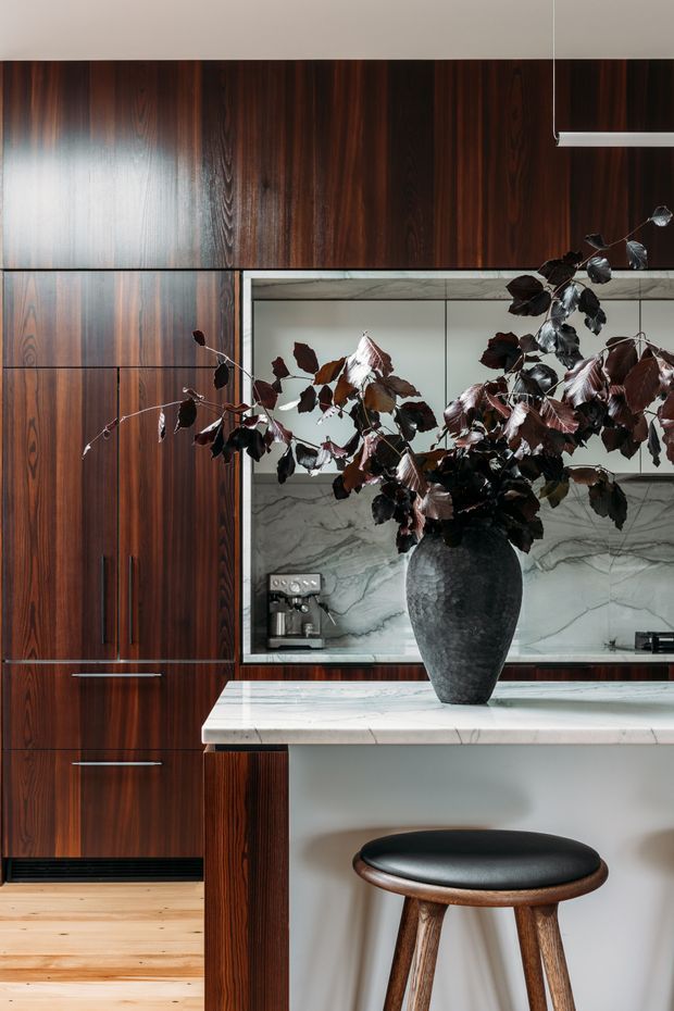 A refined contemporary kitchen featuring tall dark timber cabinetry with long integrated handles, a pale veined stone splashback and island benchtop, and a sculptural dark vase with branches. A black leather-topped timber stool sits at the island, while a minimalist linear pendant hangs above. The composition emphasizes rich wood grain, stone surfaces, and strong vertical joinery.