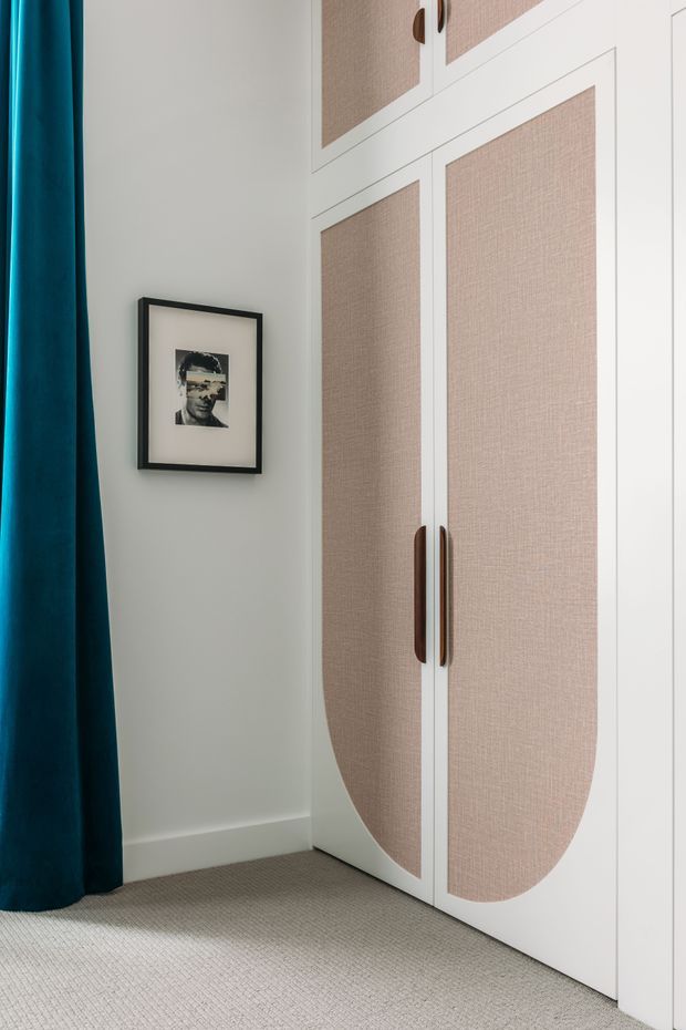 Close-up of a refined built-in wardrobe with white painted doors, inset woven beige fabric panels, and elongated dark timber handles. The wardrobe stands against a pale wall beside a deep teal floor-length curtain, with neutral carpet flooring and a small framed monochrome artwork. The image emphasizes custom joinery and soft, contemporary bedroom styling.