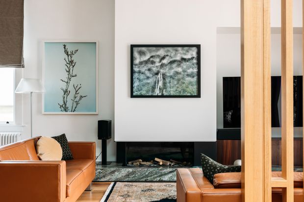 A refined contemporary living room with warm caramel leather sofas, a black-framed fireplace beneath a white feature wall, framed artwork, a patterned area rug, and a vertical timber screen or partition in the foreground. Large windows and layered neutral finishes create a bright, polished interior.