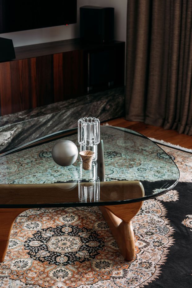 A moody, richly layered living room focused on a sculptural oval glass coffee table with a warm timber base. The table sits over an ornate patterned rug, with a dark timber media console, large television, speakers, heavy brown curtains, and a stone-like dark feature surface in the background. A small metallic sphere and clear decorative glass object provide styling accents.