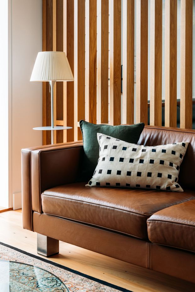 A warmly lit living room corner featuring a cognac-brown leather sofa with a matching chaise, dark green and patterned cushions, a slim floor lamp, pale timber flooring, and a vertical timber-slat screen with white backing behind the seating. A patterned area rug is partially visible in the foreground.