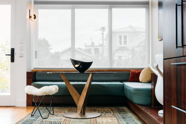 Bright, compact mid-century-inspired living room with a deep green sectional sofa beneath a wide glazed window, a sculptural wood-and-metal coffee table, patterned rug, white fur stool, and dark timber built-in cabinetry. The room has warm timber flooring, white walls, and restrained decorative accents.