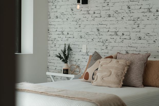 A cozy bedroom vignette with a neatly made bed layered in neutral textured cushions and a soft throw. The room features a white-painted exposed brick feature wall, a muted grey-green wall, a narrow black-framed window, a small white side table, and simple pendant lighting. Natural textures and restrained neutral tones create a relaxed, inviting atmosphere.