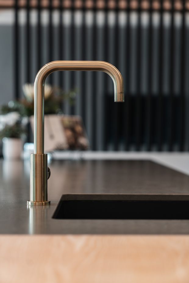 Close-up product-style photograph of a modern kitchen sink area, featuring a curved brushed brass or bronze gooseneck tap mounted on a dark benchtop beside a black inset sink. Dark vertical cabinetry or screening forms the background, with a softly blurred dining or living area beyond.