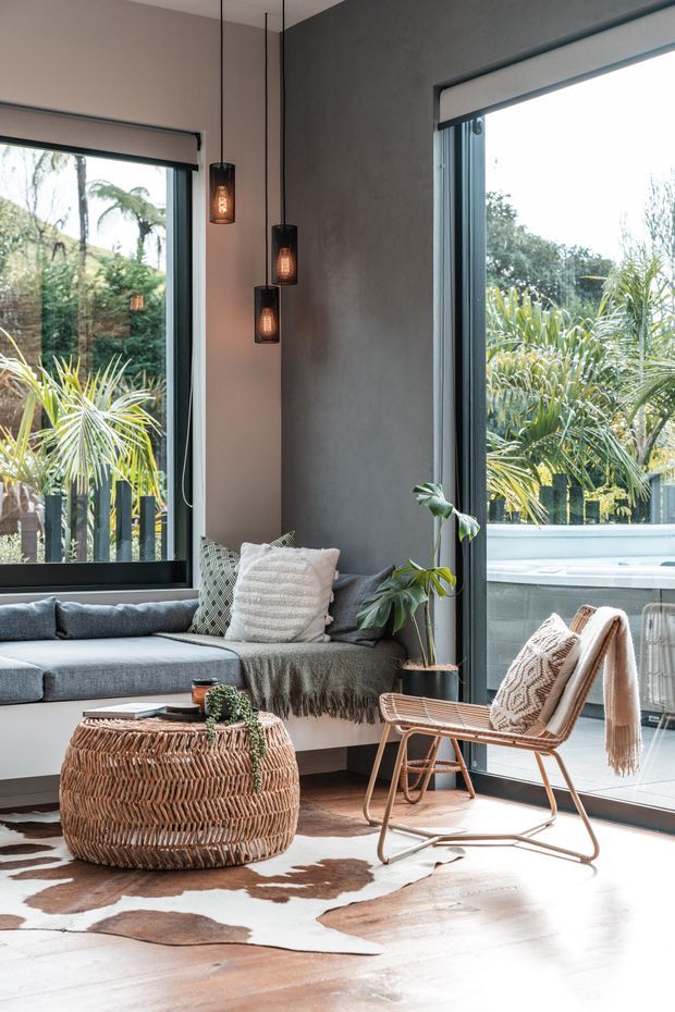 A bright, contemporary sunroom or living area with floor-to-ceiling corner glazing opening onto lush greenery. The room features a built-in grey window seat, textured cushions and throw, three suspended warm-glowing pendant lights, indoor plants, a woven wicker coffee table, a woven lounge chair, and a cowhide-style rug over a light timber floor. Dark-framed windows and a charcoal textured feature wall create contrast with the warm natural furnishings.